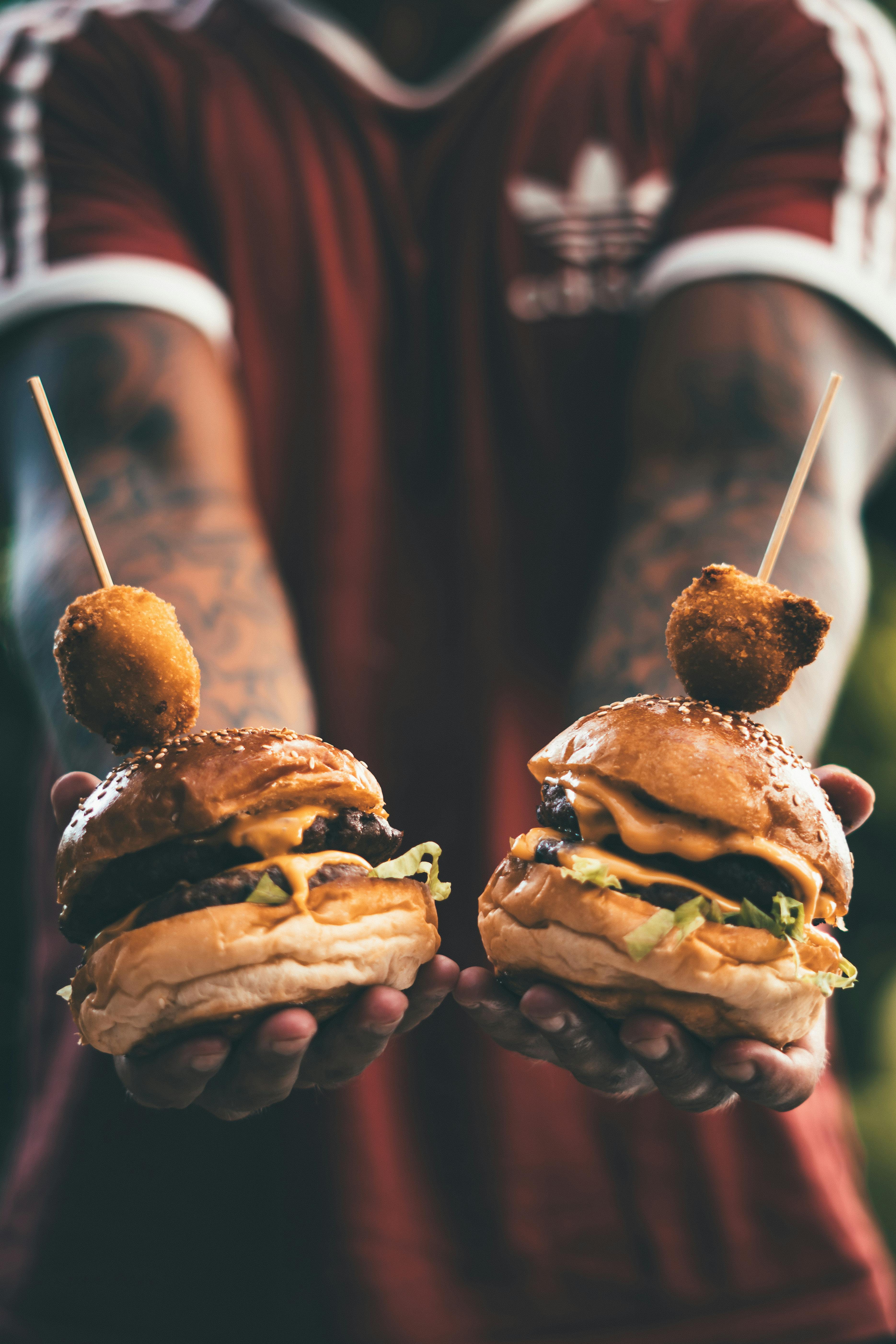Two cheeseburgers held by a tattooed individual, showcasing juicy meat, cheese, and lettuce.